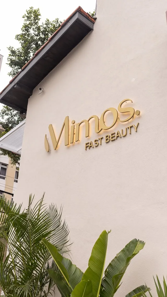 Fachada clara do salão com o letreiro dourado “Mimos Fast Beauty” e plantas tropicais na entrada.